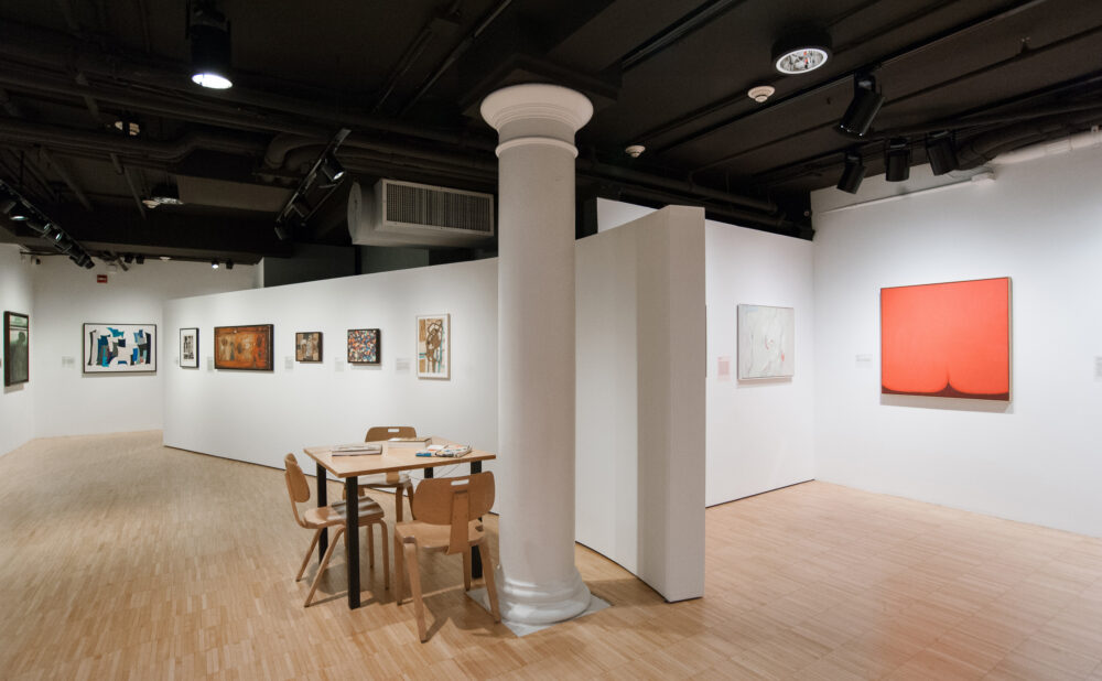 Interior of an art gallery with paintings on white walls and a table with chairs.