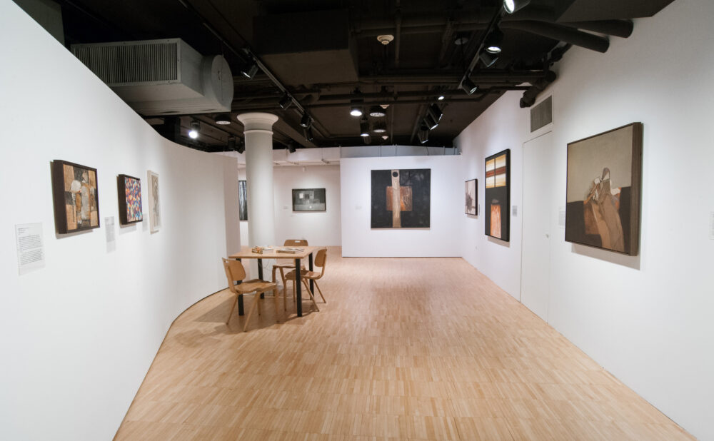 An art gallery interior with paintings on white walls and a wooden floor. A table with chairs is centered in the space.