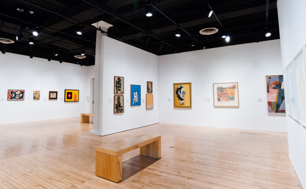 Art gallery with modern abstract paintings on white walls and wooden benches on a light wood floor.