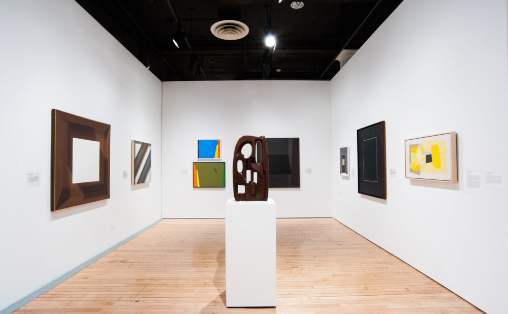 A gallery room with abstract paintings and a central wooden sculpture on a pedestal.