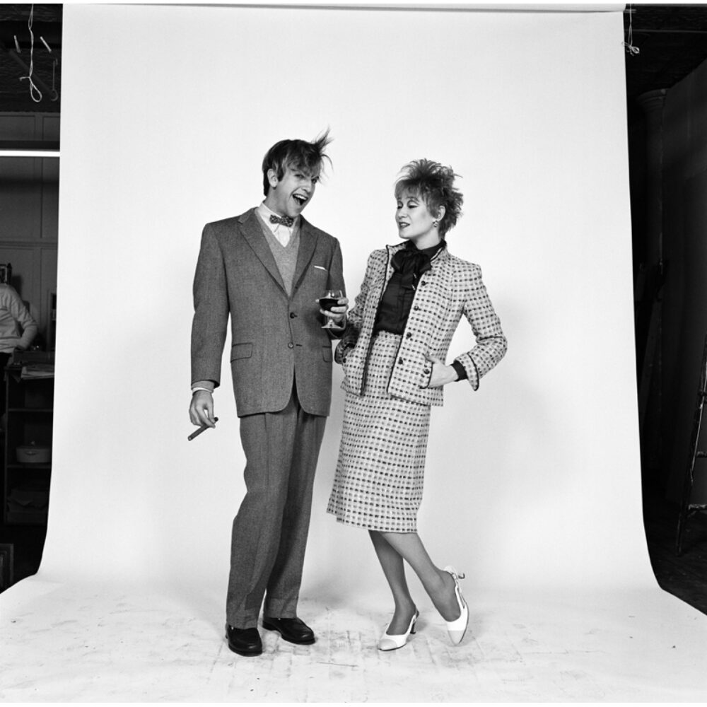 Black-and-white studio portrait photograph of Kenneth Jay Lane and a woman posing together against a white backdrop. (Alt-text generated by AI)