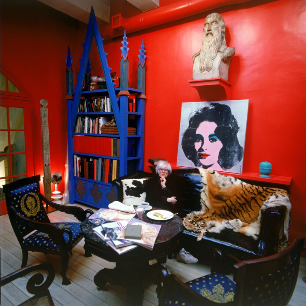 Color photograph of Andy Warhol seated in a brightly decorated room with red walls and blue shelving. (Alt-text generated by AI)
