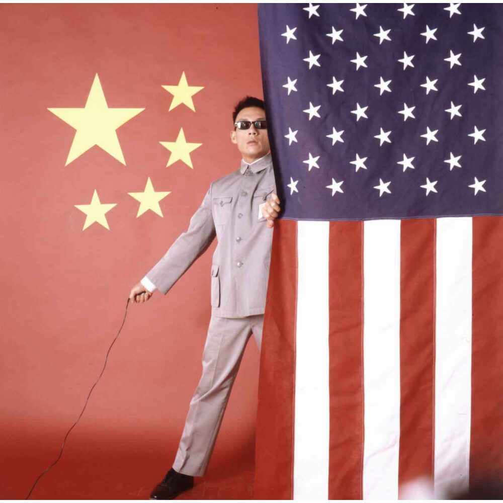 Color self-portrait of Tseng Kwong Chi in a Mao suit standing between the flag of China and the American flag. (Alt-text generated by AI)
