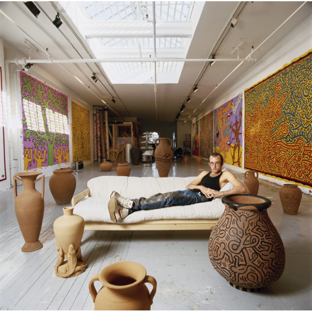 Interior portrait photograph showing Keith Haring reclining on a sofa surrounded by large ceramic vessels inside his studio. (Alt-text generated by AI)