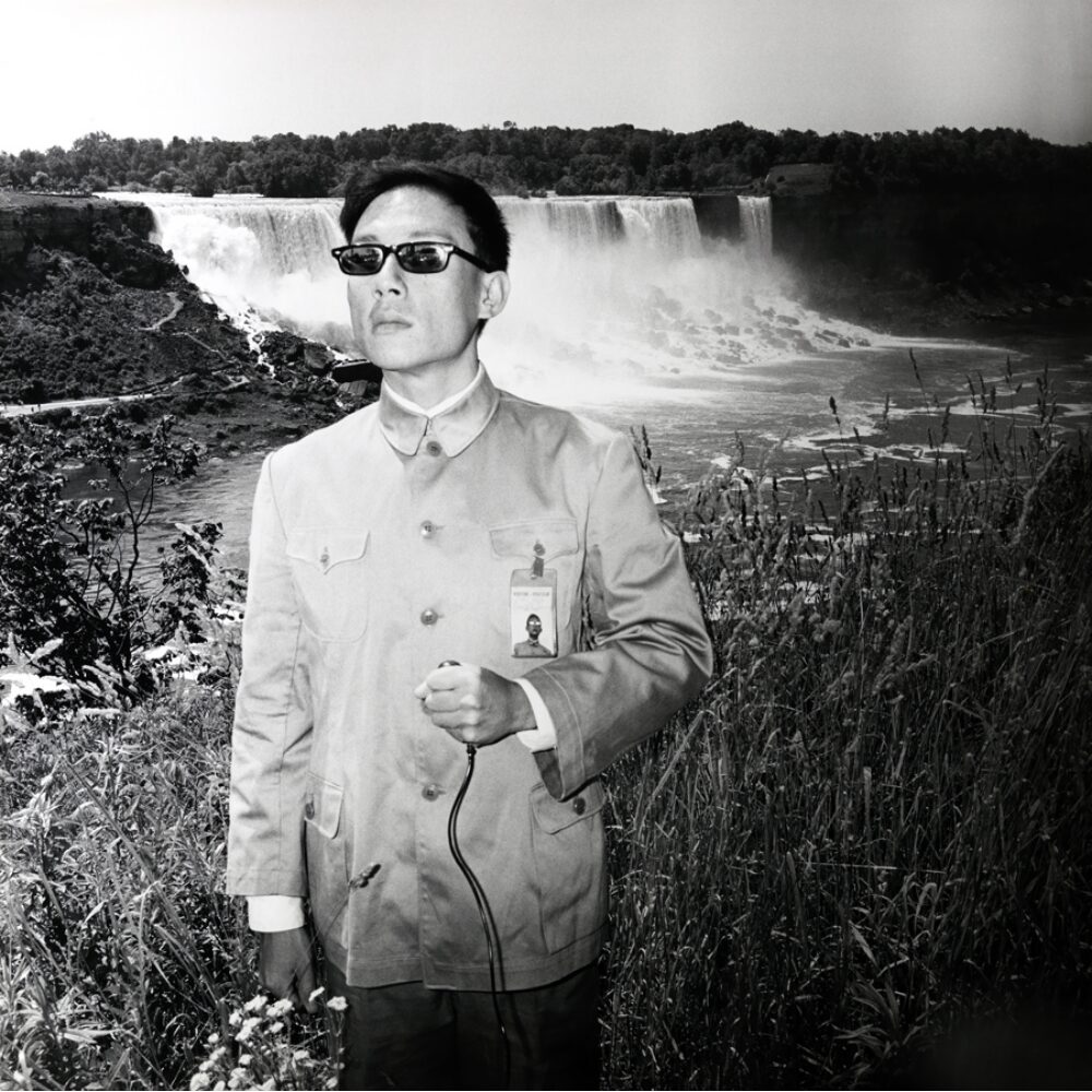 Black-and-white self-portrait of Tseng Kwong Chi wearing sunglasses and a Mao suit with Niagara Falls in the background. (Alt-text generated by AI)