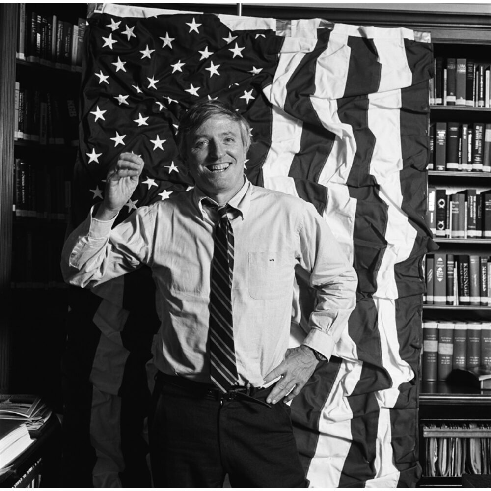 Black-and-white portrait photograph of William S. Buckley posing in front of an American flag. (Alt-text generated by AI)