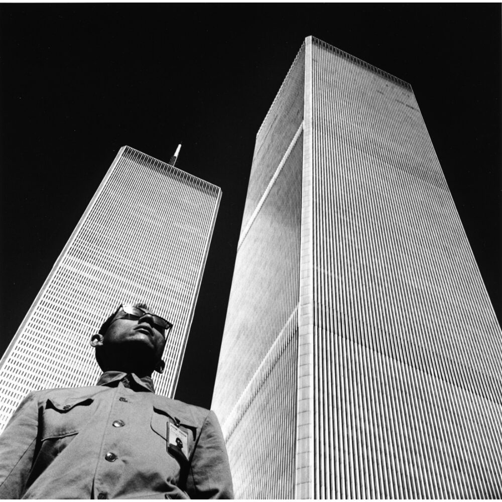 Low-angle black-and-white photograph of Tseng Kwong Chi standing in front of the Twin Towers of the World Trade Center. (Alt-text generated by AI)