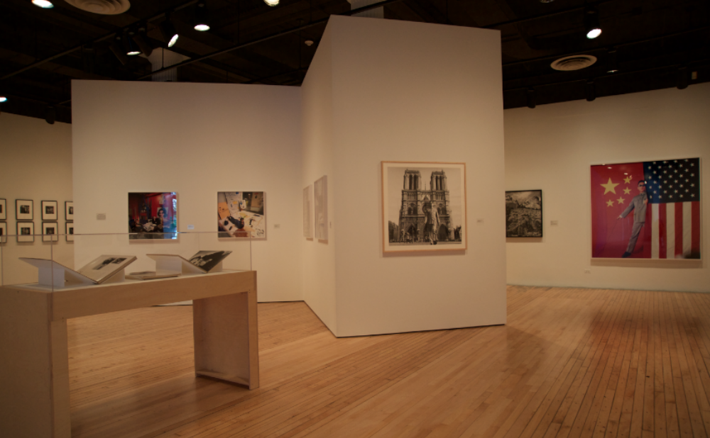 Open gallery view with a wooden vitrine on the left side, surrounded by white walls mounted with framed photographic series. (Alt-text generated by AI)