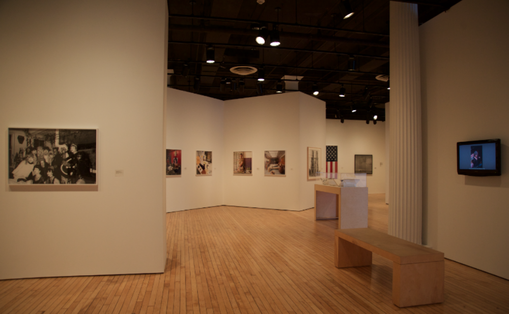 Open gallery view with white walls mounted with framed photographic series, with a wooden vitrine and a wooden bench visible on the right. (Alt-text generated by AI)