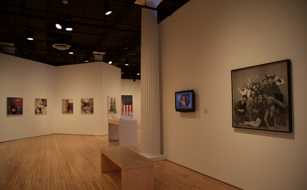 Open gallery view with white walls mounted with framed photographic series, with a wooden vitrine and a wooden bench visible on the right. (Alt-text generated by AI)