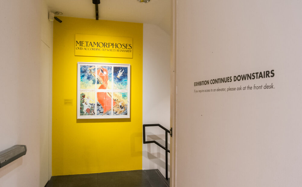 A bright yellow entryway alcove displaying a central framed painting alongside wall text next to a stairway handrail. (Alt-text generated by AI)