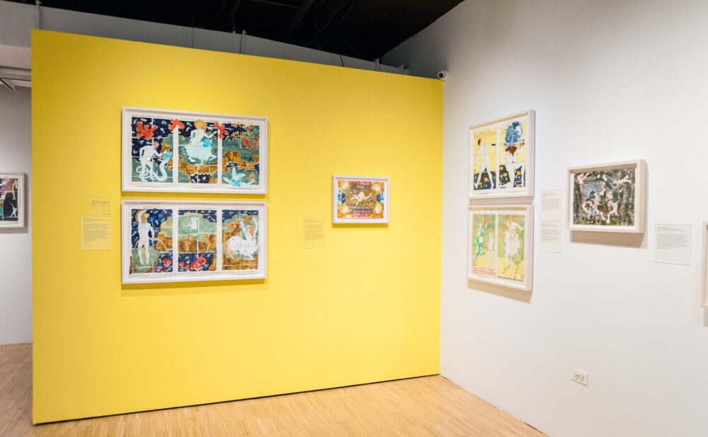 A bright yellow partition wall displaying two stacked framed artworks in an open exhibition space. (Alt-text generated by AI)