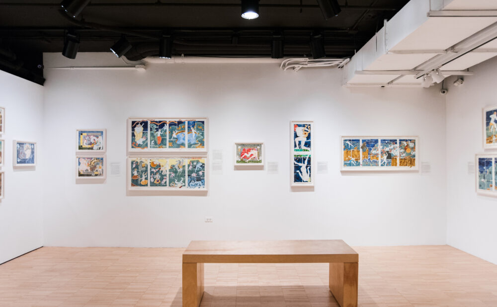 Wide view of an exhibition room featuring white walls lined with framed art and a wooden bench in the center. (Alt-text generated by AI)