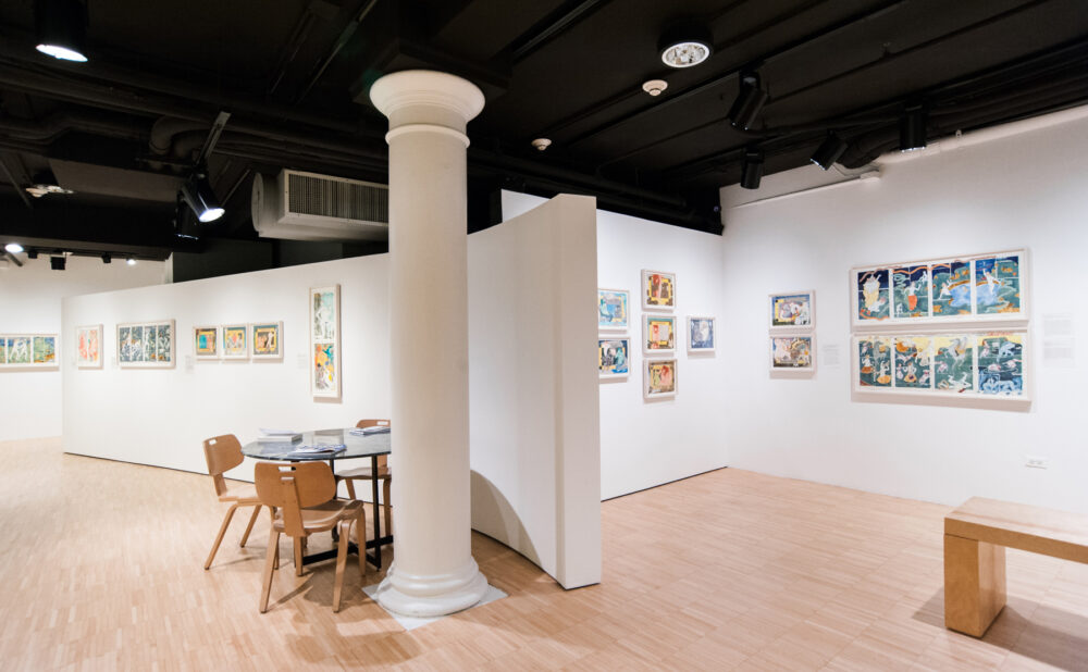 Open gallery hall with white support pillars, seating areas, and artworks mounted on surrounding walls. (Alt-text generated by AI)