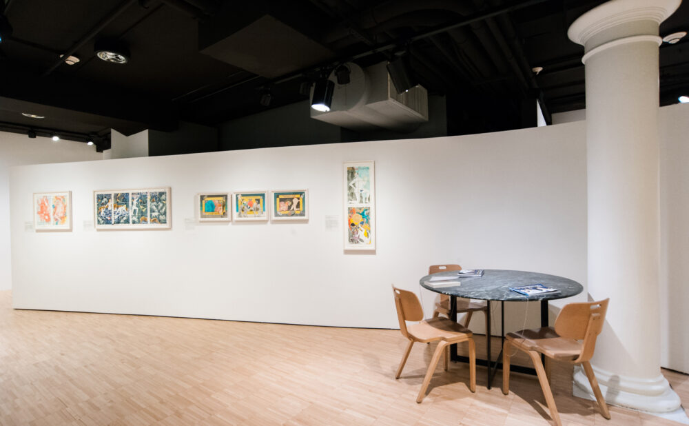 Wide exhibition view featuring a white pillar, seating table with chairs, and framed works along the back wall. (Alt-text generated by AI)