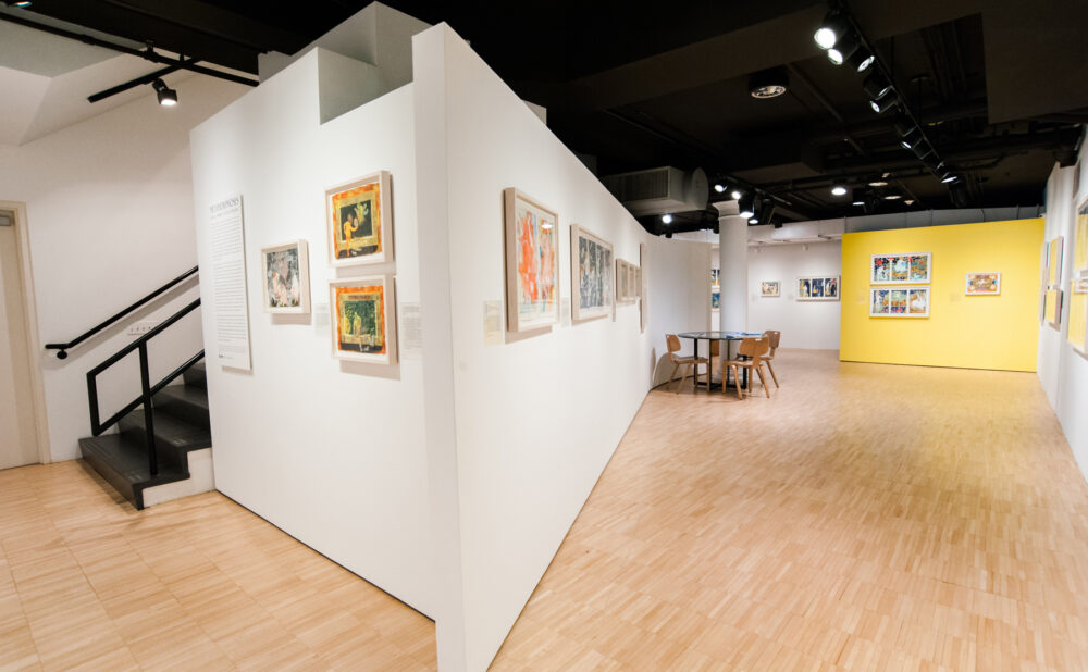 Angled view of gallery partitions leading toward a bright yellow accent wall in the distance. (Alt-text generated by AI)