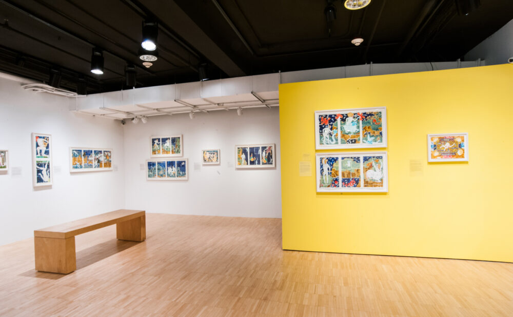 Gallery interior showing a wooden bench facing a bright yellow wall mounted with framed art. (Alt-text generated by AI)