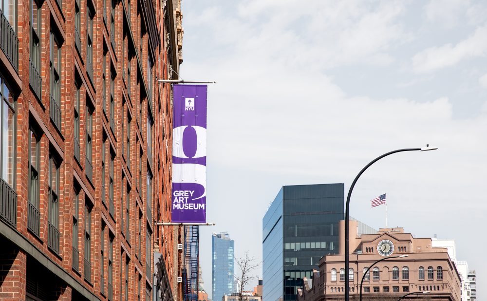 A view of downtown Manhattan above traffic on a sunny day, showing the Grey Art Museum purple banner outside of 18 Cooper Square.
