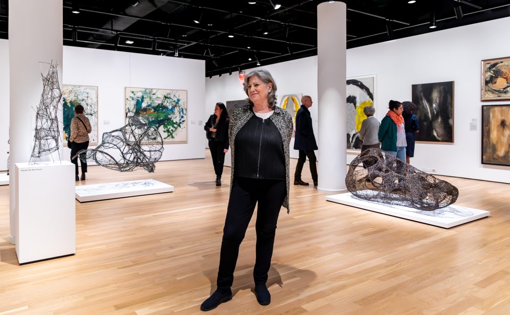 Woman with shoulder length salt and pepper hair standing in the center of an art gallery filled with sculptures and paintings. She stands with her hands behind her back, smiling away from the camera.