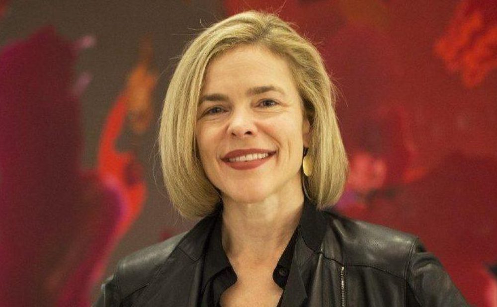 A woman with shoulder length dark blonde hair wearing a leather jacket smiles at the camera; a red, abstract, painterly background is visible.