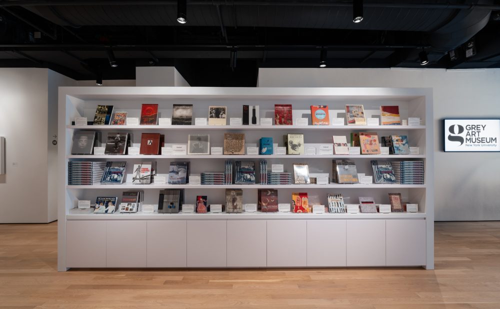 Long, white, bookshelf in an art museum, with a couple dozen artbooks on display, their covers facing the camera