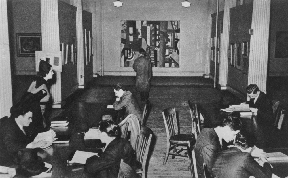 A black-and-white photo of people studying at long tables with a person looking at a large abstract painting on the back wall.