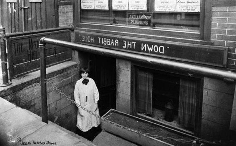 Entrance to an underground establishment named "Down the Rabbit Hole" with a woman standing at the bottom of stone stairs. The name of the establishment is written backwards, seemingly purposefully.