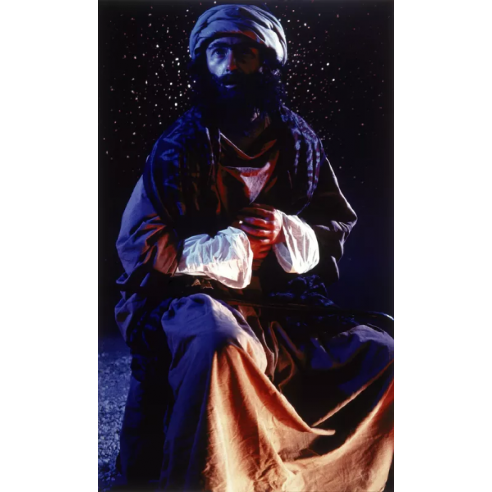 Photograph of a person seated against a dark, starry background. The person is wearing a long, flowing garment in shades of brown, with a loose-fitting head wrap. Alt-text generated by AI.