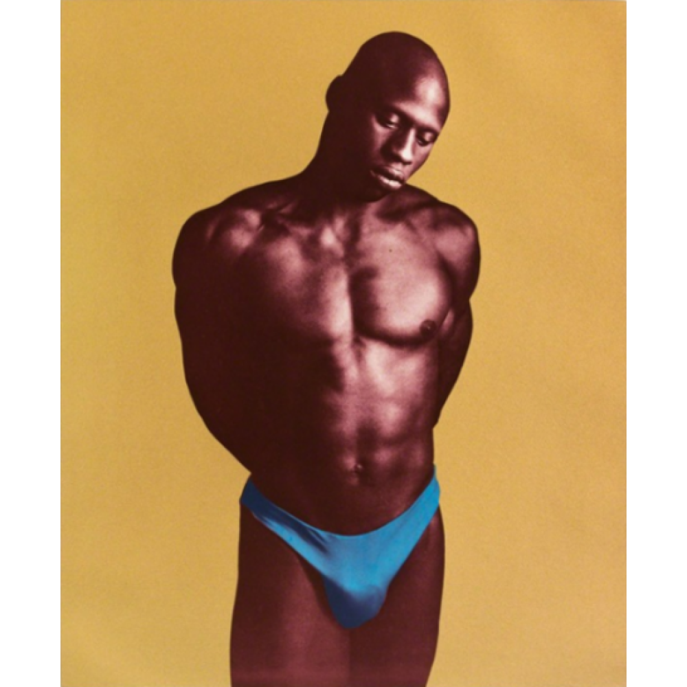 A well-muscled Black man in blue briefs stands against a mustard yellow background, gazing at the ground with his hands behind his back. Alt-text generated by AI.