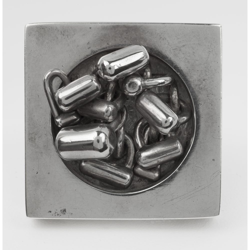 Square metal brooch with a central design of lock-like shapes.