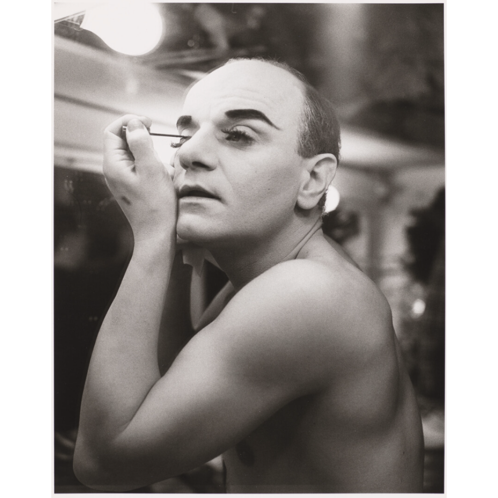 Black-and-white photograph of a shirtless balding man putting on make-up while looking in a mirror in a backstage environment.