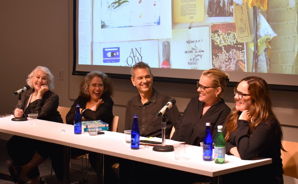 A panel of speakers seated at a long table with microphones during a presentation. Alt text generated by AI