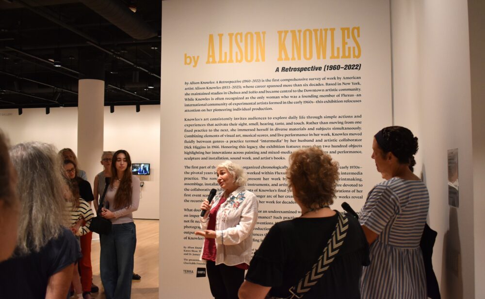 People gathered near a large wall displaying text "by Alison Knowles" inside a gallery. Alt text generated by AI