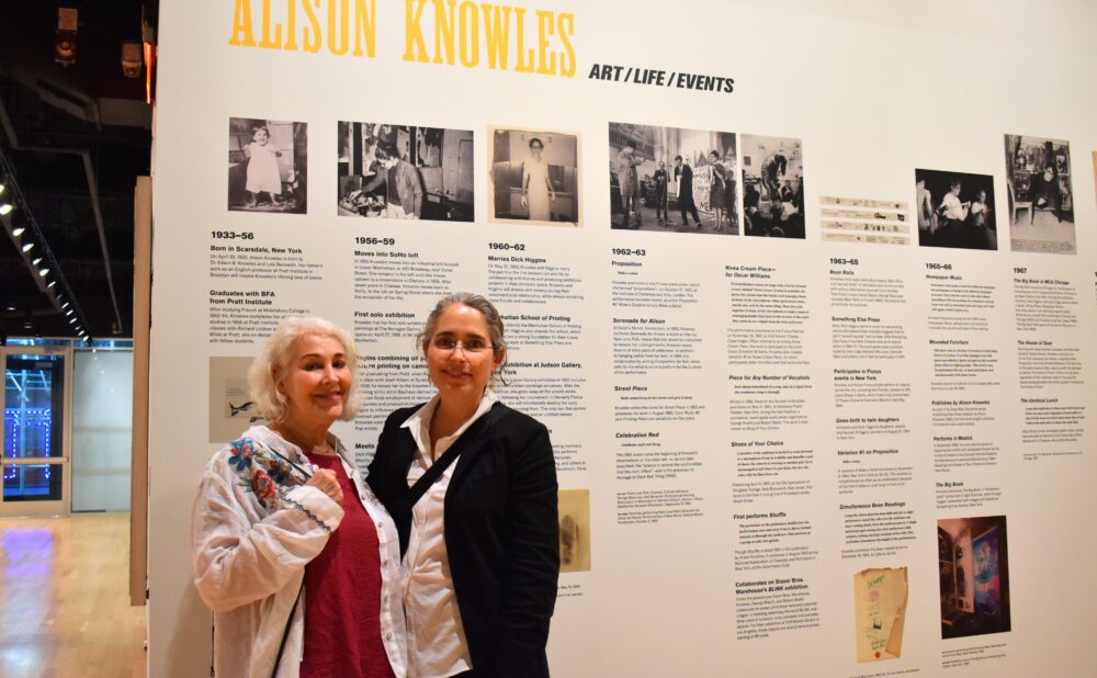 Two women standing in front of exhibition display text. Alt text generated by AI