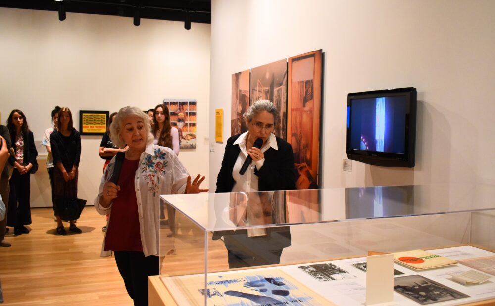 Two women standing and holding microphones in a gallery exhibition space. Alt text generated by AI