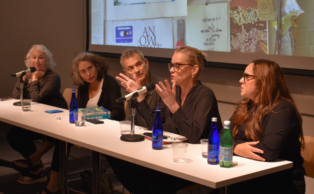 A panel of speakers seated at a long table with microphones during a presentation. Alt text generated by AI