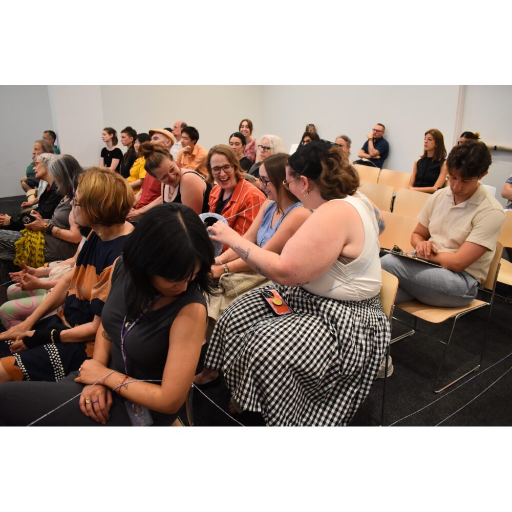 Audience members sitting in a conference room listening to a presentation and interacting with each other. Alt text generated by AI