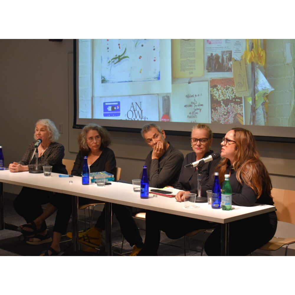 A panel of speakers seated at a long table with microphones during a presentation. Alt text generated by AI
