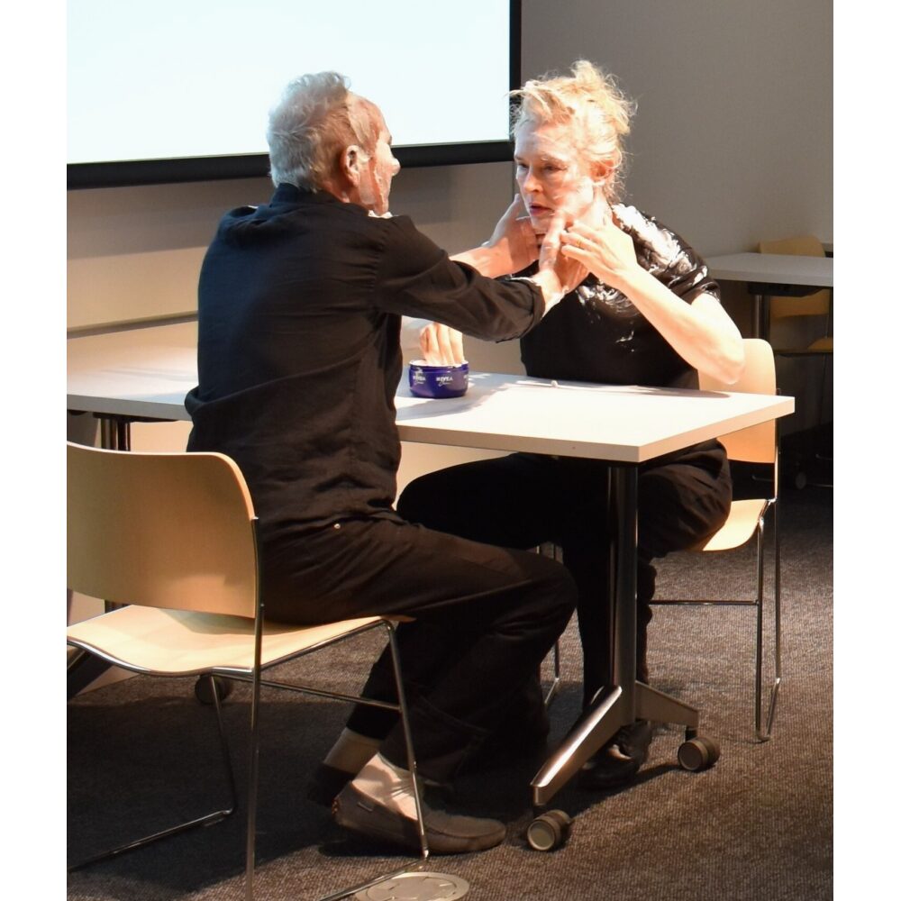 Two people seated at a small table engaging in a live reenactment of Knowles's event score, "#3: Nivea Cream Piece for Oscar Williams (1962)."