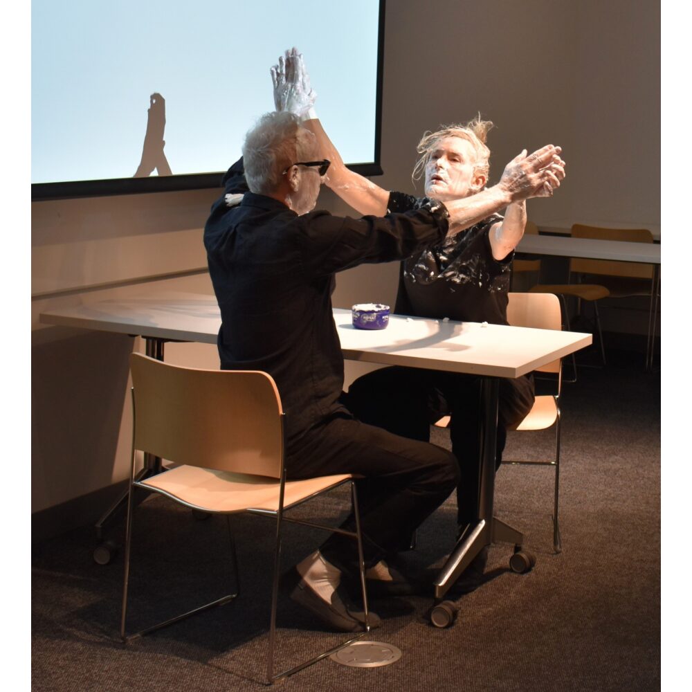 Two people seated at a small table engaging in a live reenactment of Knowles's event score, "#3: Nivea Cream Piece for Oscar Williams (1962)."