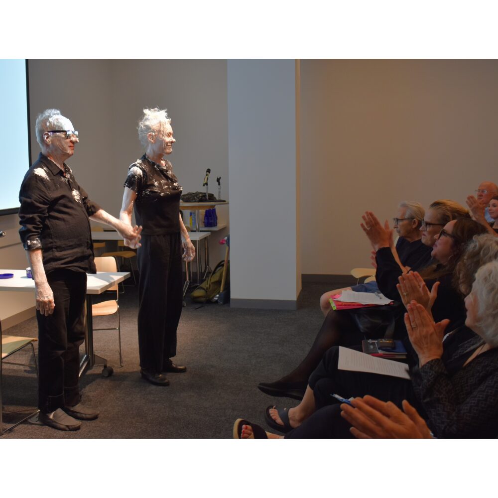 Two performers with Nivea cream on their faces, holding hands in front of an audience after their enactment of Knowles's event score "#3: Nivea Cream Piece for Oscar Williams (1962)."