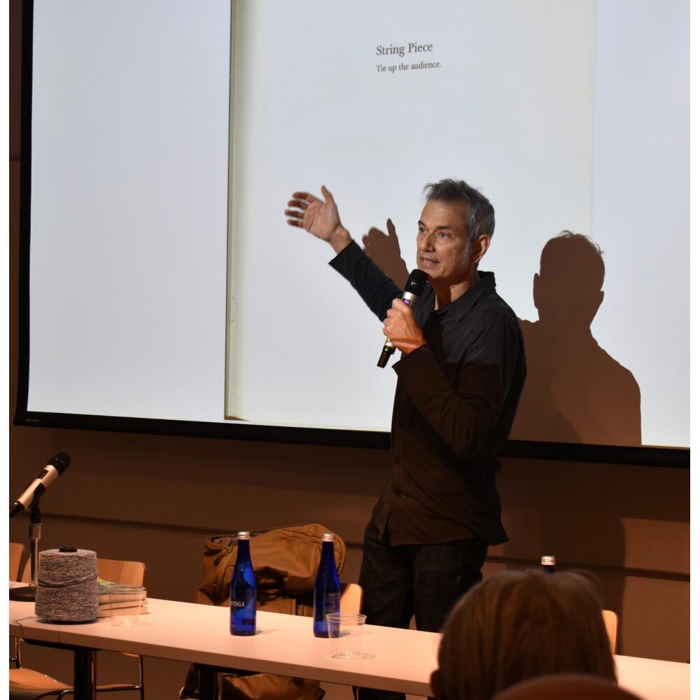 A speaker with a microphone addressing an audience in front of a screen. Alt text generated by AI