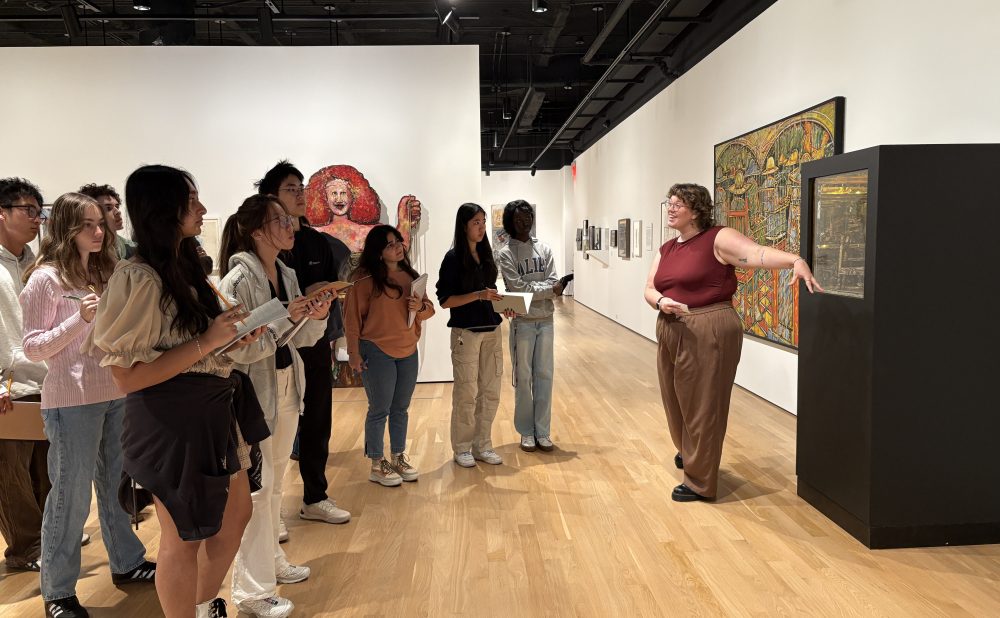 A group of people listens to a tour guide in an art gallery. Alt-text generated by AI.