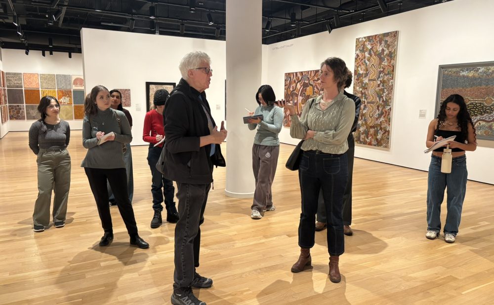 People in an art gallery, with two in the foreground conversing, surrounded by Aboriginal paintings. Alt-text generated by AI.