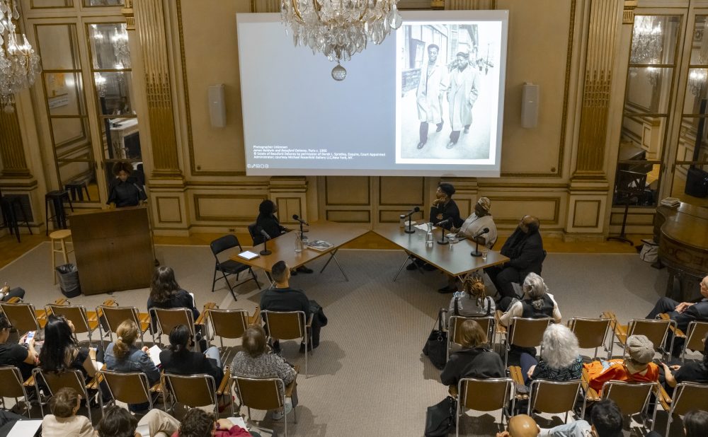 A presentation in an ornate room with chandeliers, showing a black and white photo on a large screen. Audience members sit in rows facing a podium and panelists. Alt-text generated by AI.