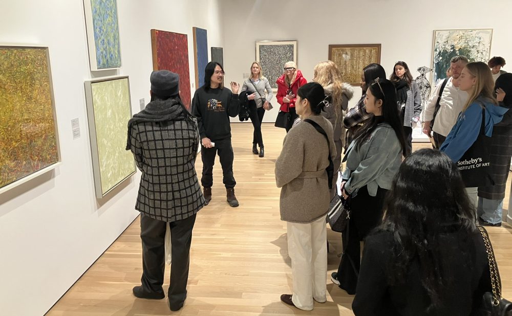 People observing art on a gallery wall during a guided tour.  Alt-text generated by AI.