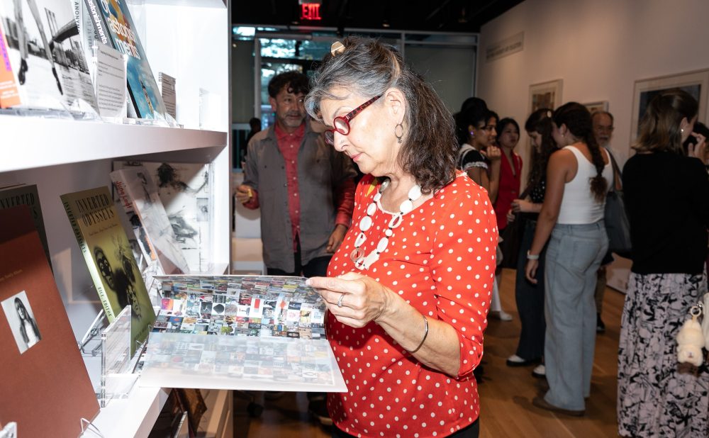 A woman in a red polka dot shirt looks at a book in a gallery with others mingling around. Alt-text generated by AI.