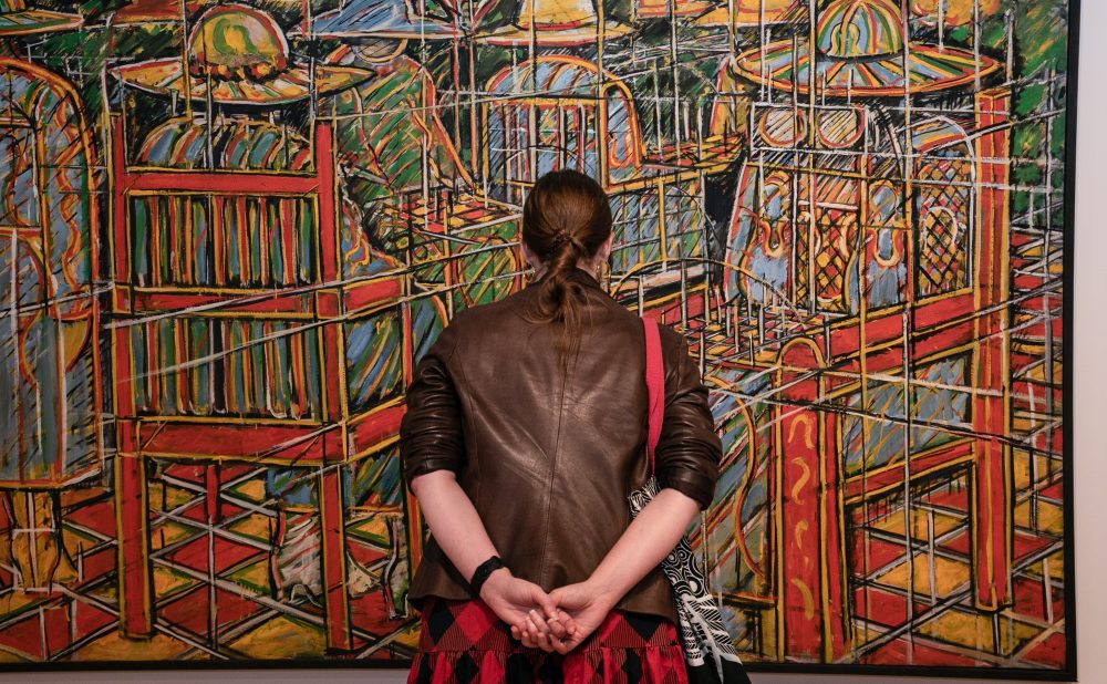A person in a brown leather jacket and patterned skirt stands observing a colorful abstract painting. Alt-text generated by AI.