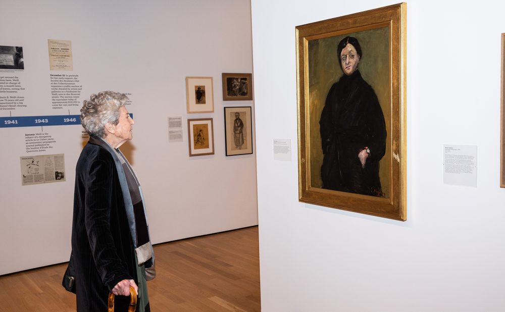 Elderly woman with a cane observes a framed portrait in an art gallery, surrounded by smaller artworks on a white wall.