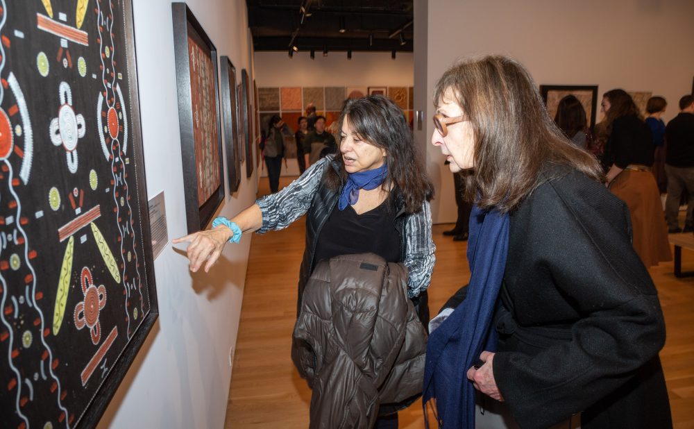 Two women examining an artwork in a crowded gallery. Alt-text generated by AI.